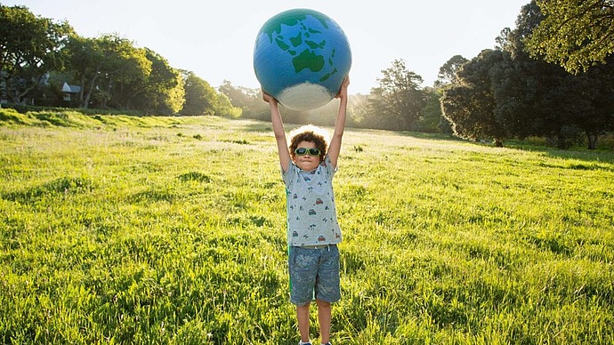 Ein Junge mit Sonnenbrille und Sommerkleidung steht auf einer sonnigen Wiese und hält eine große aufblasbare Weltkugel über seinen Kopf, während im Hintergrund Bäume und Grünflächen zu sehen sind.