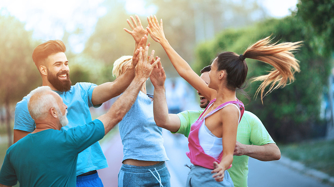 Eine Gruppe von fünf Personen unterschiedlichen Alters, in legerer Sportkleidung, geben sich im Freien fröhlich High Fives. Sie wirken voller Energie und glücklich, umgeben von viel Grün an einem sonnigen Tag.